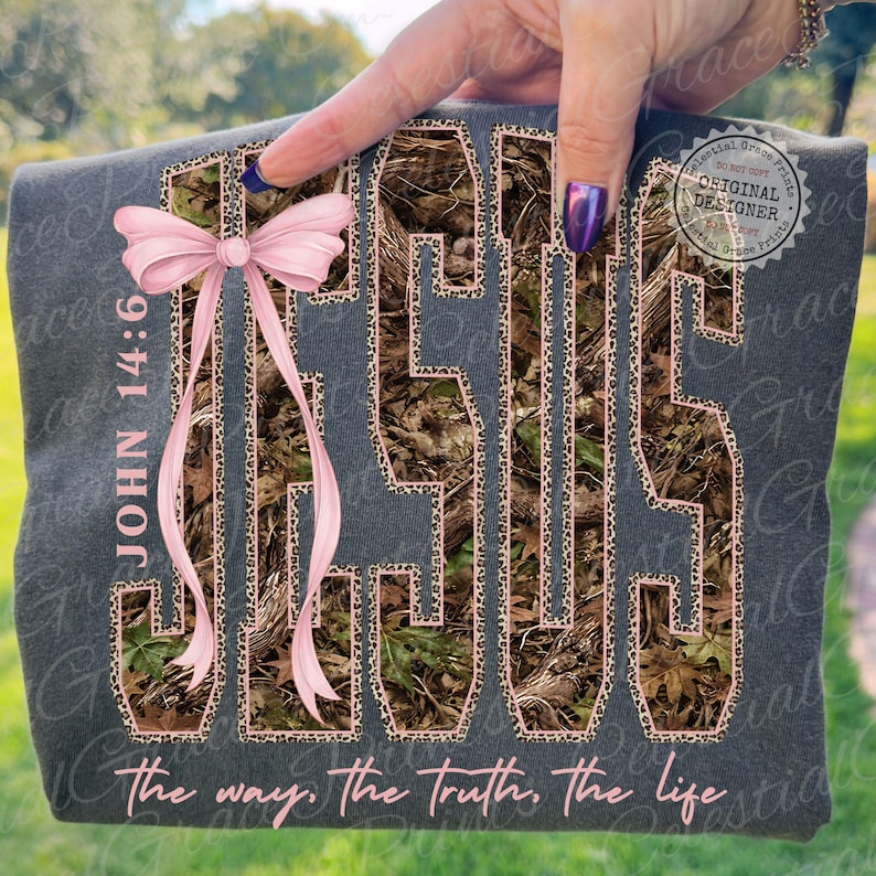 May include: Dark gray t-shirt with the word "JESUS" in a camouflage pattern outlined in leopard print. A pink bow is tied to the left side. The text "JOHN 14:6" and "the way, the truth, the life" are also visible.