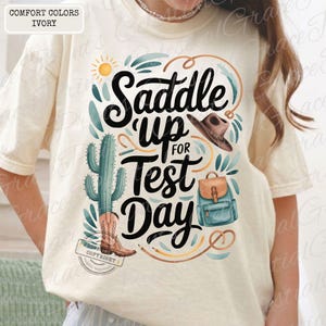 May include: Ivory-colored t-shirt with a graphic that says "Saddle Up for Test Day." The design includes a cactus, cowboy hat, backpack, and boots. The shirt is a comfort color.