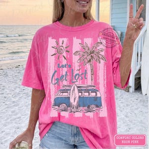 May include: Neon pink t-shirt with a beach-themed graphic. The design includes a sun, palm tree, a blue van with a surfboard, and the text "Let's Get Lost". The shirt is a Comfort Colors brand.