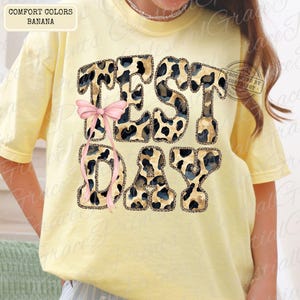 May include: Yellow t-shirt with the words "TEST DAY" in a leopard print design, accented with a pink bow. The shirt is a comfort color in the shade of banana.
