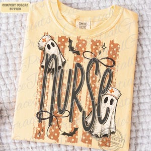 May include: A butter yellow t-shirt featuring a graphic print with the word NURSE in large black script. The design includes two white ghost figures wearing nurse caps with orange crosses, orange vertical stripes, and small black bats.