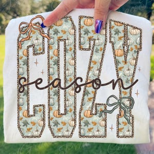 May include: White long-sleeved shirt with the words "COZY SEASON" in a fall-themed design. The letters are filled with pumpkins and leaves, outlined with a leopard print. Decorative bows and stars accent the design.