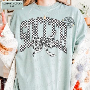 May include: A light blue t-shirt with the word "SOCCER" in a checkered pattern. A soccer ball bow is centered below the word. The shirt has the text "COMFORT COLORS BAY" in the upper left corner.