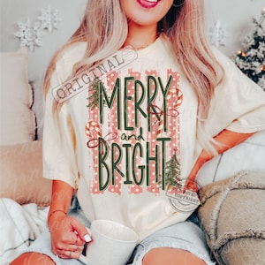 May include: A cream-colored short-sleeve t-shirt featuring a graphic print with vertical red and white stripes. The text reads MERRY and BRIGHT in dark green lettering, accompanied by illustrations of two candy canes and two pine trees.