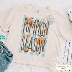 May include: An ivory short-sleeve t-shirt featuring a graphic print with the text PUMPKIN SEASON in dark brown letters. The design includes orange pumpkin illustrations, light blue vertical stripes with yellow dots, and thin orange line accents.