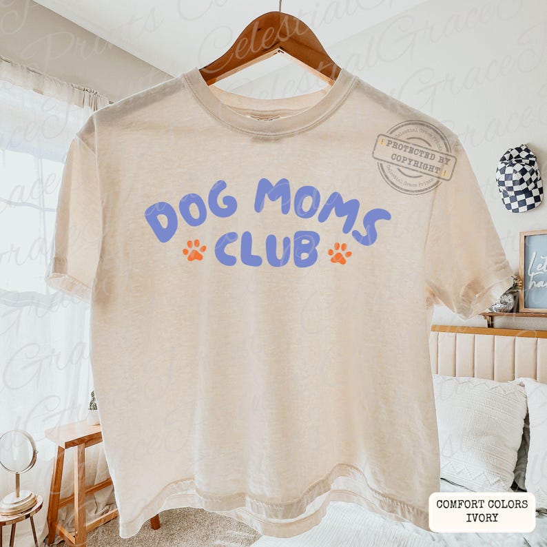 May include: Ivory-colored t-shirt with the text "DOG MOMS CLUB" in blue, featuring orange paw prints. The shirt is hanging on a wooden hanger. The bottom right corner of the shirt has the text "COMFORT COLORS IVORY".