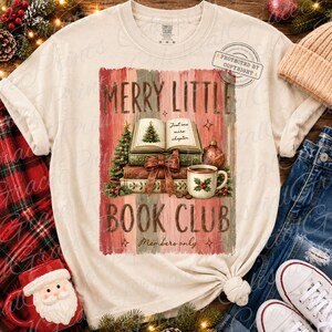May include: A cream-colored short-sleeve t-shirt featuring a graphic of a stack of books, a Christmas tree, and a mug. The text reads MERRY LITTLE BOOK CLUB, Just one more chapter, and Members only. The design includes red and green holiday accents.