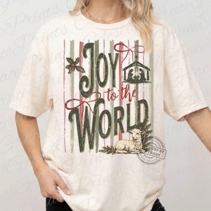 Puede incluir: Una camiseta de manga corta color crema con un gr&aacute;fico de rayas verticales en rojo y verde apagados. El texto Joy to the World aparece en cursiva verde oscuro. El dise&ntilde;o incluye un bel&eacute;n, acebo y la ilustraci&oacute;n de un peque&ntilde;o cordero.