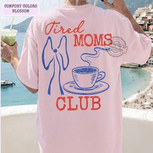 May include: A light pink t-shirt with the words "Tired Moms Club" in red and blue. The design includes a blue bow, a coffee cup, and a saucer. The shirt is a comfort colors blossom.