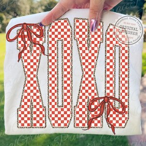 May include: White t-shirt featuring the word "MOM" in large, bold letters. The letters have a red and white checkered pattern with a leopard print outline. Red bows are placed on the top left and bottom right of the shirt.