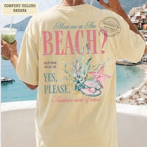 May include: A cream-coloured t-shirt with the words "Meet me at the BEACH?" in pink and blue. The back of the shirt features a seashell, starfish, and aquatic plant graphic. The words "Salty Hair, Ocean Air, Yes, Please, Sunshine state of mind" are also printed.
