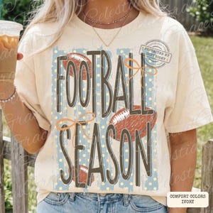 May include: An ivory short-sleeve t-shirt featuring a graphic print with the text FOOTBALL SEASON in dark brown letters. The design includes three brown football illustrations, light blue vertical stripes, and orange line-drawn bows.