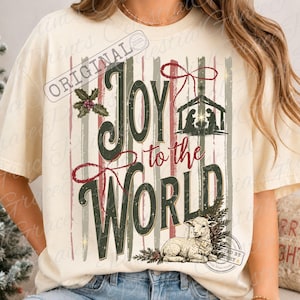 Puede incluir: Una camiseta de color crema con un gr&aacute;fico que incluye el texto Joy to the World en verde oscuro y rojo. El dise&ntilde;o presenta una silueta de bel&eacute;n, un cordero, hojas de acebo y rayas verticales en tonos rojos y verdes apagados.