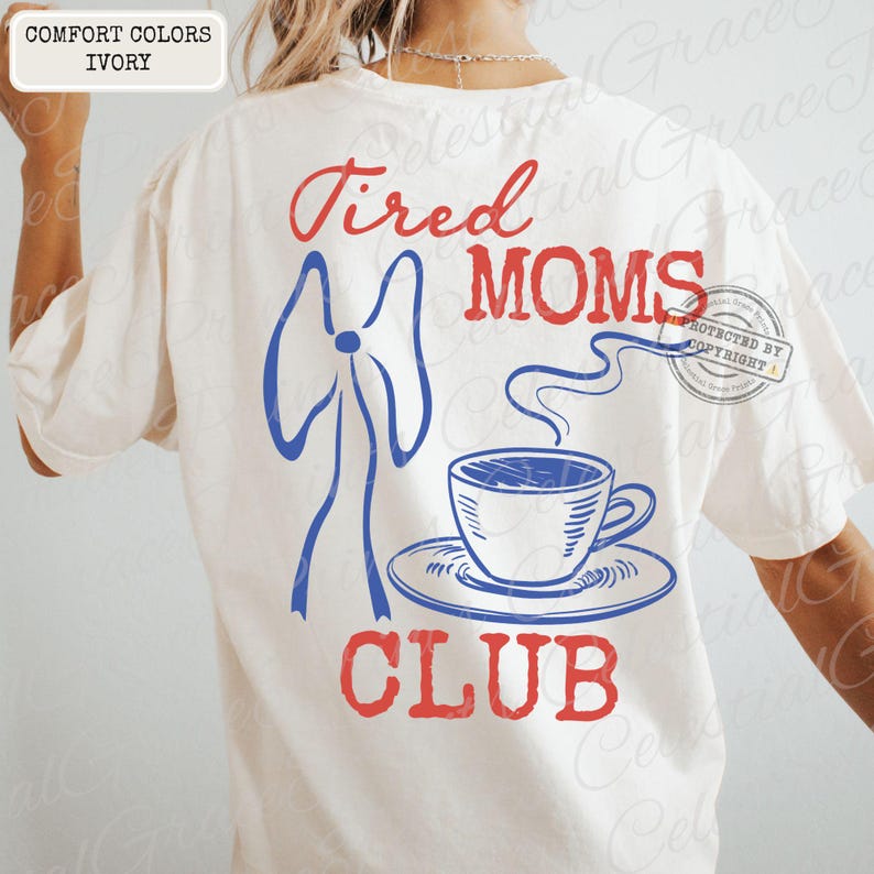 May include: Ivory-colored t-shirt with the words "Tired Moms Club" in red and blue. A blue bow and a steaming coffee cup graphic are also featured. The shirt is a comfort color.