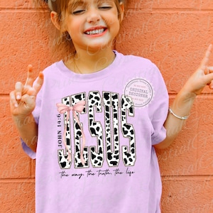 Puede incluir: Una camiseta de color lila claro con la palabra "JESUS" en un dise&ntilde;o de estampado de vaca y un lazo rosa. El texto "JOHN 14:6" est&aacute; impreso verticalmente a la izquierda. La frase "the way, the truth, the life" est&aacute; impresa debajo.
