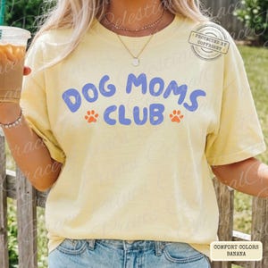 May include: A light yellow t-shirt with the text "DOG MOMS CLUB" in blue, with orange paw prints. The shirt is a Comfort Colors brand, and the color is Banana.