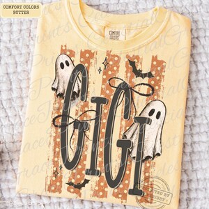 May include: A butter yellow short-sleeve t-shirt featuring the text GIGI in large black letters. The design includes two white ghosts, black bats, and small star shapes over a background of vertical orange stripes with white polka dots.