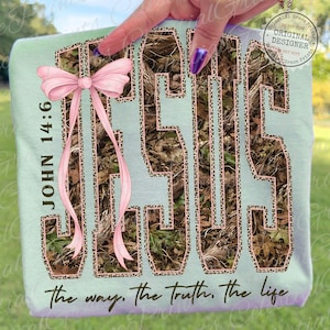 May include: A light blue t-shirt with the word "JESUS" in a camouflage pattern with a leopard print outline. The shirt has a pink bow and the text "JOHN 14:6" and "the way, the truth, the life."