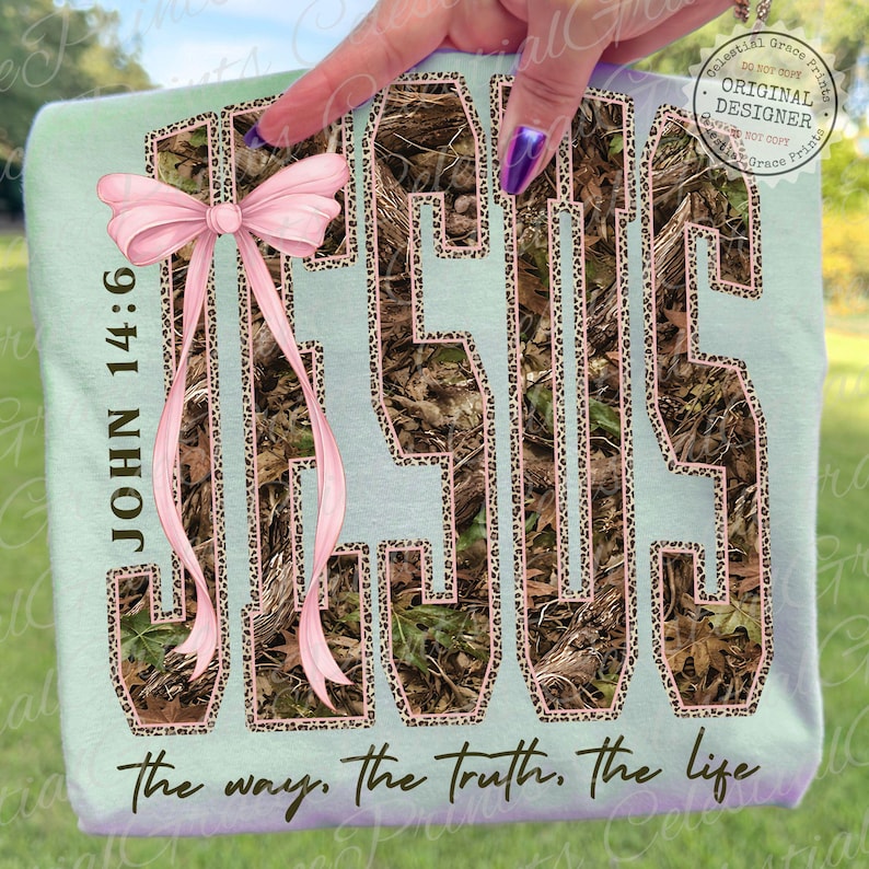 May include: A light blue t-shirt with the word "JESUS" in a camouflage pattern with a leopard print outline. The shirt has a pink bow and the text "JOHN 14:6" and "the way, the truth, the life."