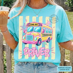 May include: A lagoon blue t-shirt featuring a graphic of a yellow school bus with the text SCHOOL BUS and DRIVER in pink. The design includes vertical yellow stripes and pink and blue star accents. The shirt is made of cotton fabric.