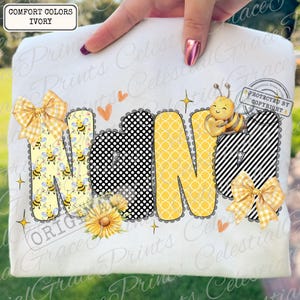 May include: Ivory t-shirt featuring the word "NANA" in a colourful design. The letters are decorated with bee and floral patterns, polka dots, and stripes. It includes bows, a sunflower, and a cute bee graphic.