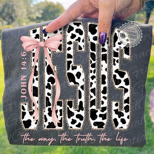Puede incluir: Camiseta gris oscuro con la palabra "JESUS" en un dise&ntilde;o de estampado de vaca y borde de estampado de leopardo. Un lazo rosa adorna el lado izquierdo. El texto "JOHN 14:6" y "the way, the truth, the life" tambi&eacute;n son visibles.