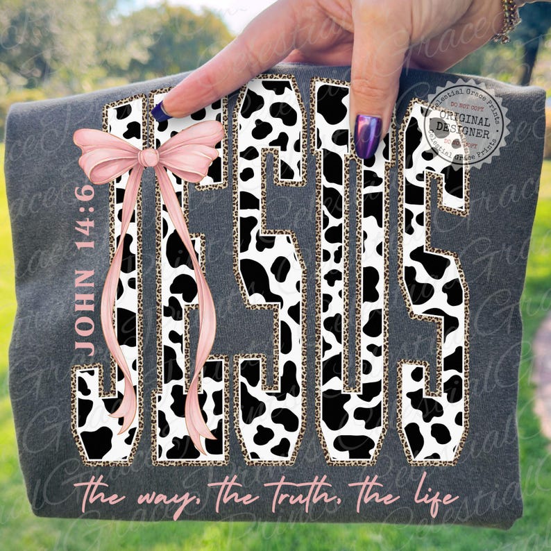 Puede incluir: Camiseta gris oscuro con la palabra "JESUS" en un dise&ntilde;o de estampado de vaca y borde de estampado de leopardo. Un lazo rosa adorna el lado izquierdo. El texto "JOHN 14:6" y "the way, the truth, the life" tambi&eacute;n son visibles.