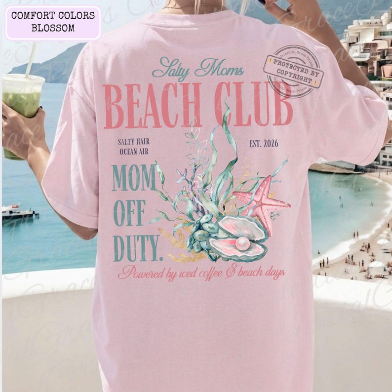 Puede incluir: Camiseta rosa claro con la frase "Salty Moms Beach Club" en un estilo vintage. La parte trasera de la camiseta presenta un dise&ntilde;o de concha, estrella de mar y algas marinas. El texto "Mom Off Duty" tambi&eacute;n est&aacute; impreso en la camiseta.