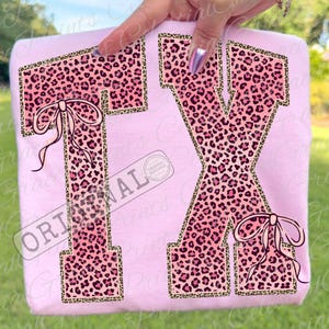 May include: A light pink sweatshirt featuring the letters "TX" in a pink leopard print design with a gold border. The letter "T" has a bow on the left side, and the letter "X" has a bow on the bottom right. The word "ORIGINAL" is printed on the bottom left.