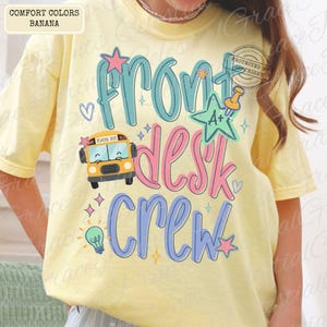 Könnte beinhalten: Ein hellgelbes T-Shirt mit dem Schriftzug "Front Desk Crew" in Türkis, Rosa und Blau. Das Design umfasst einen Schulbus, Sterne, eine Glühbirne und eine Reißzwecke. Das Shirt ist mit "Comfort Colors Banana" gekennzeichnet.