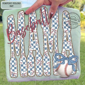 May include: A light green t-shirt with the words "Baseball Mama" in a distressed, red script. The letters are filled with a blue and white checkered pattern and baseball-themed designs. A baseball with a blue bow is at the bottom.