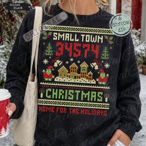 May include: A black long-sleeve sweatshirt featuring a festive knit-style graphic with the text SMALL TOWN 34574, CHRISTMAS, and HOME FOR THE HOLIDAYS. The design includes gingerbread houses, Christmas trees, bells, and two small figures.