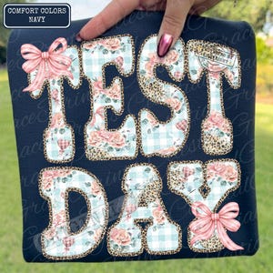 May include: Navy blue t-shirt with the words "TEST DAY" in a decorative font. The letters are adorned with floral, gingham, and leopard print patterns, and accented with pink bows. The top left corner of the shirt has the text "COMFORT COLORS NAVY".