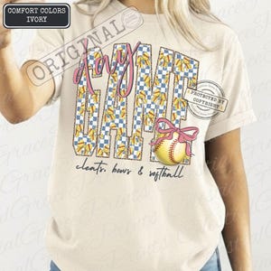May include: Ivory Comfort Colors t-shirt with a softball theme. The word "Day" is in pink script, and "Game" is in a blue and white checkered pattern with yellow bows. A softball with a pink bow is on the right. The text "cleats, bows & softball" is at the bottom.