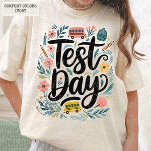 May include: Ivory-colored t-shirt with a floral design and the words "Test Day" in large black script. The design includes pink and yellow flowers, school buses, pencils, and a backpack. The shirt is made of Comfort Colors material.