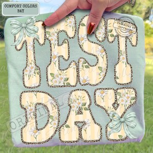 Peut inclure: Un t-shirt vert clair avec les mots "TEST DAY" en grandes lettres stylis&eacute;es. Les lettres ont un fond ray&eacute; jaune et blanc avec des motifs floraux de marguerites et des bordures en imprim&eacute; l&eacute;opard. Le t-shirt a deux n&oelig;uds vert clair.
