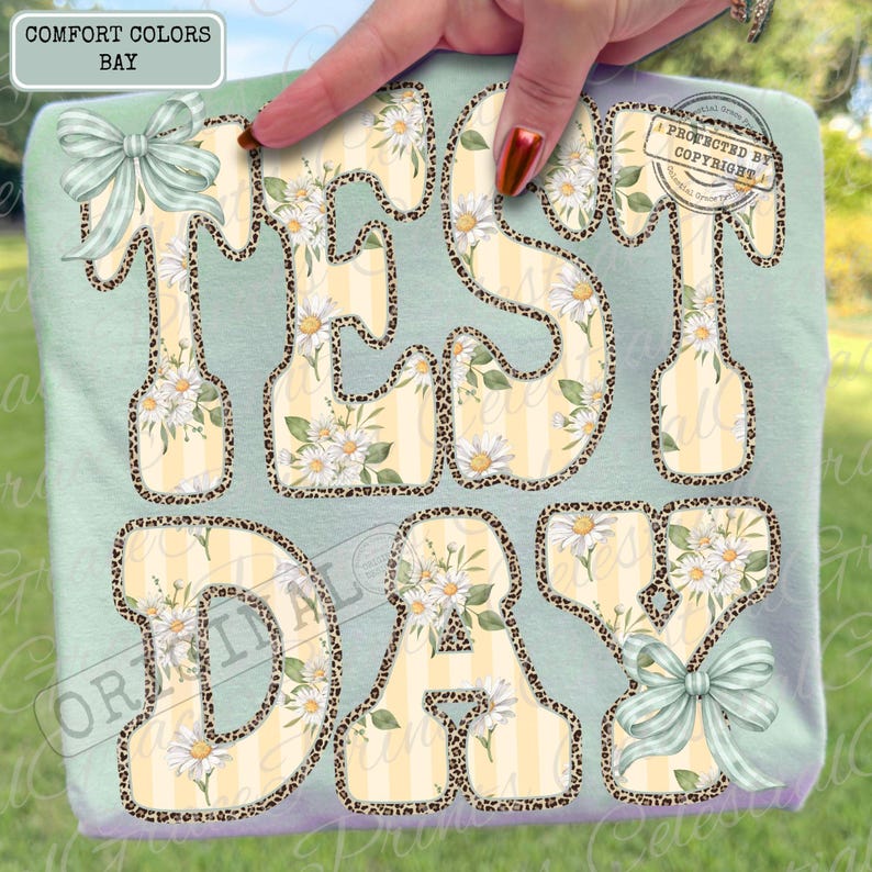 Peut inclure: Un t-shirt vert clair avec les mots "TEST DAY" en grandes lettres stylis&eacute;es. Les lettres ont un fond ray&eacute; jaune et blanc avec des motifs floraux de marguerites et des bordures en imprim&eacute; l&eacute;opard. Le t-shirt a deux n&oelig;uds vert clair.