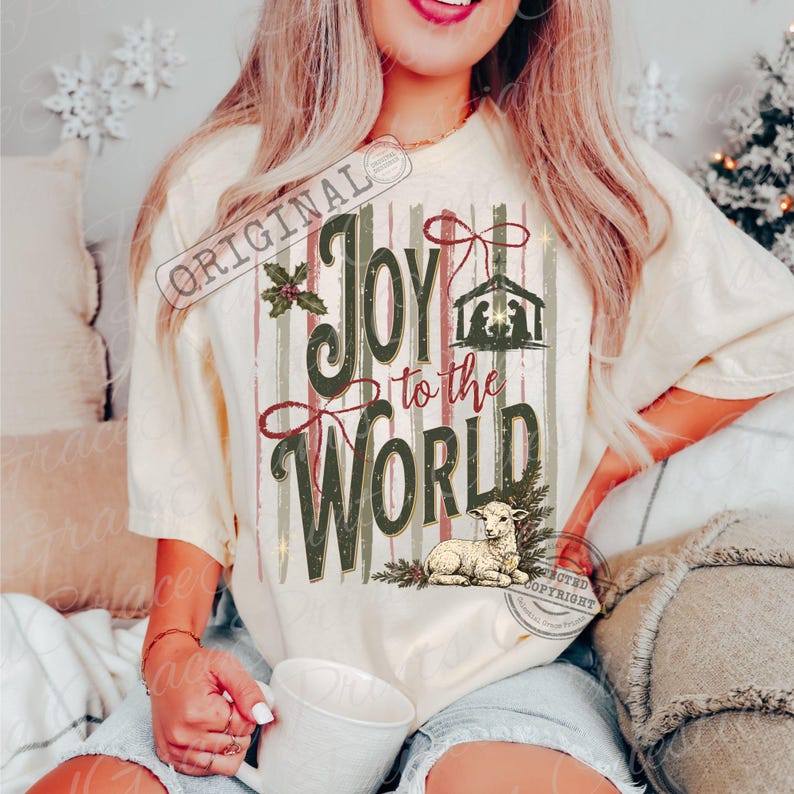 Puede incluir: Una camiseta de manga corta color crema con un estampado gr&aacute;fico que incluye el texto Joy to the World. El dise&ntilde;o presenta una silueta de bel&eacute;n, un lazo rojo, hojas de acebo y una ilustraci&oacute;n de un cordero sobre rayas verticales en tonos suaves.
