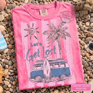 May include: Neon pink t-shirt with a graphic of a blue and white van carrying surfboards, a palm tree, and the text "Let's Get Lost." The design includes a sun and birds, evoking a beach theme.