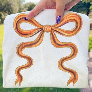 May include: A white sweatshirt featuring a large, detailed illustration of a bow. The bow is rendered in shades of orange, gold, and brown, with a textured appearance. The design is centered on the fabric.