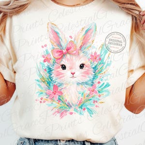 May include: Cream-colored t-shirt featuring a watercolor illustration of a bunny with a pink bow, surrounded by flowers and teal accents. The bunny has gold glitter accents on its ears. The text "Celestial Grace Prints" is printed on the shirt.