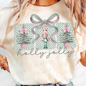 May include: Cream-coloured t-shirt with a Christmas design. The design features a nutcracker, Christmas trees, and a bow, in pink, green, and white. The words "holly jolly" are written in a script font below the design.