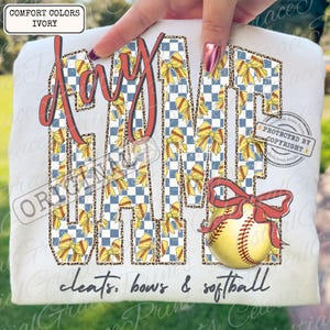 May include: Ivory Comfort Colors t-shirt with the words "Day Game" in a red, bow-filled design. A softball with a red bow is on the bottom right. The text "cleats, bows & softball" is at the bottom.