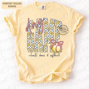 May include: A light yellow t-shirt with the words "Game Day" in a pink and checkered design. A softball with a pink bow is featured below the text, with the words "cleats, bows & softball" below the softball.