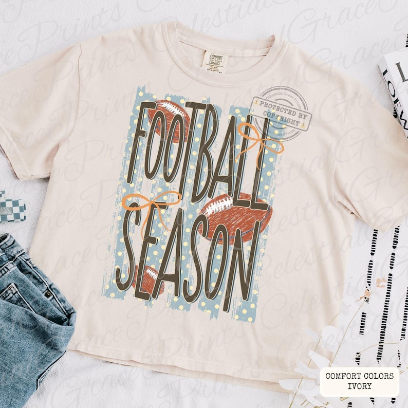 May include: An ivory short-sleeve t-shirt featuring the text FOOTBALL SEASON in dark brown letters. The design includes a light blue background with yellow polka dots, three brown football illustrations, and orange line-drawn bows.