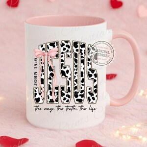 Puede incluir: Taza de cer&aacute;mica blanca con interior y asa rosas. La palabra "JESUS" est&aacute; impresa en la parte delantera con un dise&ntilde;o de estampado de vaca en blanco y negro, con un lazo rosa. Tambi&eacute;n se ve el texto "JOHN 14:6" y "the way, the truth, the life".