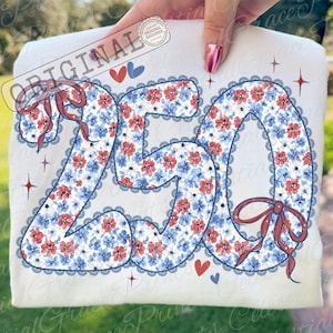 Op de afbeelding: Een wit t-shirt met het nummer "250" in een bloemenpatroon van rode en blauwe bloemen. De cijfers zijn omlijnd met een blauwe kanten rand en versierd met rode strikken, harten en sterren. Het woord "ORIGINAL" staat linksboven.