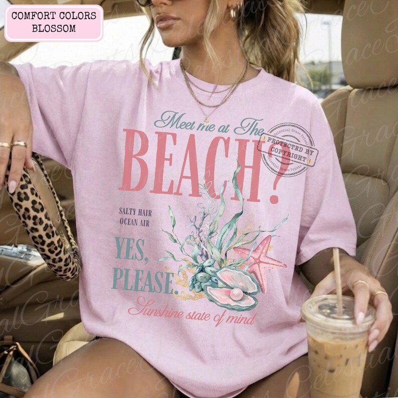 May include: A pale pink t-shirt with the words "Meet me at The BEACH?" in a retro style. The shirt has a seashell and starfish design with the phrases "Salty Hair, Ocean Air, Yes, Please, Sunshine state of mind."