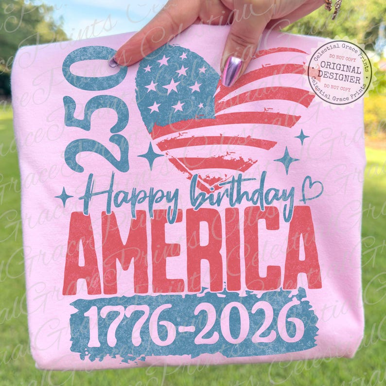 Puede incluir: Una camiseta rosa claro con un dise&ntilde;o patri&oacute;tico. El dise&ntilde;o incluye una bandera estadounidense en forma de coraz&oacute;n, el texto "Happy Birthday AMERICA", los n&uacute;meros "250" y "1776-2026". La camiseta est&aacute; sostenida por una persona.