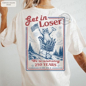 May include: Ivory-colored t-shirt with a graphic featuring a raccoon in a shopping cart, waving an American flag and holding a hot dog. The text reads "Get in Loser" and "We're celebrating 250 YEARS EST. 1776".